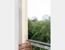 Blick von einem Balkon mit Holzstuhl und Metallgeländer auf eine grüne Baumlandschaft, im Hintergrund mehrere hohe Wohngebäude.