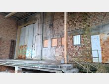 Verwitterte Laderampe eines alten Industriegebäudes mit großer, rostiger Doppeltür und gemauerten Ziegelwänden mit abblätterndem Putz. Mehrere kleine, teils vernagelte Fenster, ein Metallgeländer an einer Betontreppe sowie sichtbare Rohre und Leitungen prägen die Fassade.