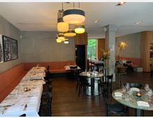 Innenraum eines modernen Restaurants mit warmem, minimalistischen Design. Holzboden, graue Betonwände, orangegepolsterte Sitzbänke, gedeckte Tische mit weißem Tischtuch, schwarzen Stühlen und dekorativer Beleuchtung.