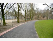 Ein öffentlicher Park mit kahlen Bäumen im Winter, einem geteerten Weg, der eine Wiese umrahmt, und einem umzäunten Sandplatz im Hintergrund. Im Hintergrund sind mehrstöckige Gebäude zu erkennen.