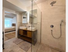 Modernes Badezimmer mit begehbarer Glasdusche, beige gefliesten Wänden und zwei weißen Waschbecken auf einer Holzkommode. Im Hintergrund ist ein angrenzendes Schlafzimmer mit einem Bett und Holzfußboden zu sehen.