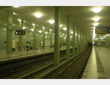 U-Bahn-Station mit grünen Säulen, glasierten Wandfliesen und einem gewölbten Deckenbereich, der mit runden Leuchten ausgestattet ist. Gleise führen durch die Station, während Personen auf den Bahnsteigen warten; gut sichtbar sind Beschilderungen, Uhr und Werbeplakate.