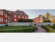 Mehrstöckige rote Wohngebäude mit Satteldächern und weißen Fenstern, umgeben von einer gepflegten Grünanlage mit Hecken und kleinen Bäumen. Ein gepflasterter Weg führt durch den Garten, vorbei an einer niedrigen Steinmauer.