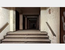 Blick von einer kurzen Treppe in einen verlassenen, langen Flur mit abblätternden Wänden, Handläufen und mehreren Stützenpfeilern. Der Raum wirkt staubig und dunkel, mit niedriger Decke und einem fernen Durchgang am Ende des Korridors.