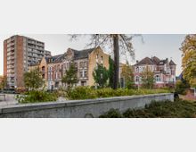 Städtische Szenerie mit historischen Gebäuden im Vordergrund, darunter reich verzierte Altbauten und eine rosa Villa. Im Hintergrund steht ein moderner, mehrstöckiger Wohnblock, umgeben von Bäumen und Grünanlagen.