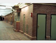 Ein unterirdischer Korridor mit Backsteinwänden, beige-grünen Türen und beleuchteten Schildern. Der Boden ist mit Fliesen in geometrischen Mustern gestaltet, und die Decke verfügt über abgehängte Lampen.