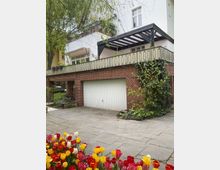 Mehrfamilienhaus mit weißer Fassade, Garage im Erdgeschoss mit rotem Klinker umgeben, darüber eine große Terrasse mit Geländer und Pergola. Im Vordergrund sind bunte Tulpenbeete und ein gepflasterter Weg zu sehen.