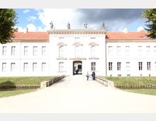 Helles, barockes Schlossgebäude mit symmetrischer Fassade, roten Ziegeldächern und Statuen auf dem Dach. Ein Weg über eine kleine Brücke führt zum zentralen Torbogen, durch den Personen gehen, mit Rasenflächen und Bäumen im Vordergrund.