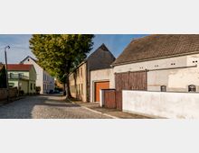 Straßenszene in einem ruhigen Dorf mit Kopfsteinpflaster, alten Gebäuden mit teils verputzten Fassaden und roten Ziegeldächern; rechts ein größerer Scheunenbau mit Holztoren, links moderne Wohnhäuser und ein großer Baum.