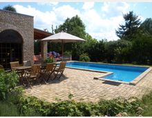 Ein Garten mit gepflasterter Terrasse, einem rechteckigen Pool, einer Sitzgruppe aus Holzstühlen und Tisch unter einem Sonnenschirm. Im Hintergrund befinden sich ein Backsteingebäude mit großen Rundbogenfenstern und üppige grüne Vegetation.