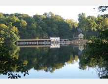 Ein See mit ruhiger Wasseroberfläche, die die umliegenden Bäume und Gebäude spiegelt. Am Ufer befinden sich ein Bootsverleih unter einer überdachten Halle sowie ein kleines gelbes Haus, umgeben von dichter Vegetation.