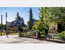 Wohnstraße in einem Vorort mit Einfamilienhäusern, umgeben von gepflegten Gärten. Im Vordergrund ist ein Zaun mit Bepflanzung, im Hintergrund ein Haus mit Walmdach und großen Fenstern, umgeben von Bäumen und Straßenlaternen.