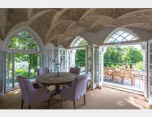 Eleganter Innenraum mit dekorativem Gewölbedecken und großen Rundbogenfenstern, die auf eine Terrasse mit Holztisch und -stühlen führen; im Vordergrund steht ein runder Holztisch mit lilafarbenen Polsterstühlen. Durch die Fenster sind grüne Bäume und ein Haus im Garten sichtbar.