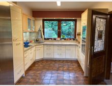 Moderne Küchenzeile mit hellen Holzfronten und granitfarbenen Arbeitsplatten, zentralem Fenster mit Blick auf einen grünen Garten, braunen Fliesenboden und gläserner Küchentür mit geometrischem Muster.