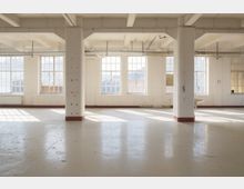 Heller, leerer Industrie-Loftraum mit breiten Stützpfeilern, sichtbaren Deckenbalken und großen Sprossenfenstern entlang der Außenwand. Der glatte Boden reflektiert das einfallende Tageslicht; Leitungen und Heizkörper sind an Decke und Wand sichtbar.