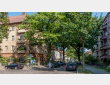 Straßenszene in einem Wohnviertel mit mehrstöckigen Gebäuden im Altbaustil, Balkonen und großen Fenstern. Die Straße ist von Bäumen gesäumt, teilweise mit parkenden Autos, und eine Person schiebt ein Fahrrad am Gehweg entlang.