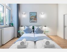 Helles, modern eingerichtetes Schlafzimmer mit zwei zusammengestellten Einzelbetten, grauen Kissen und gerollten Handtüchern am Fußende. Neben großen Fenstern mit Vorhang stehen Glas-Nachttische mit Lampen; an der Wand hängen ein Berlin-Poster und zwei Wandleuchten.