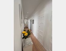 Schmaler, heller Flur mit Holzdielenboden, weißen Wänden und Türen. Im Vordergrund steht ein kleiner Marmortisch mit einer Vase gelber Blumen, im Hintergrund sind eine geöffnete Tür und ein Spiegel sichtbar.