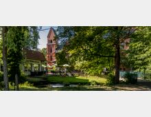 Ein kleiner Park mit einem Bachlauf, Bäumen und Grünflächen, im Hintergrund eine rote Backsteinkirche mit einem spitzen Turm. Links ein Café mit einer Terrasse und Sonnenschirmen, rechts eine Fußgängerbrücke.