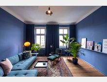 Wohnzimmer mit blauen Wänden, Holzboden und einer Einrichtung im modernen Stil. Zu sehen sind ein blaues Sofa, ein Sessel, Zimmerpflanzen, ein persischer Teppich, große Fenster mit dunklen Vorhängen und dekorative Bilder auf einer niedrigen Wandablage.