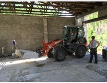 Ein offenes, unfertiges Gebäude mit Sichtbetonwänden, in dem sich ein Kompaktlader der Marke "Atlas" befindet, der einen Betonteil transportiert. Zwei Personen stehen bzw. sitzen im Arbeitsbereich, während Sonnenlicht durch ein unvollständiges Dachgestell einfällt.