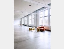 Ein modernes, helles Loft mit hohen Decken, großflächigen Fenstern und grauem Betonboden. Im Vordergrund eine Sitzecke mit einem orangefarbenen Sessel, gelben Hockern und einem Glastisch, im Hintergrund minimalistische Möbel.