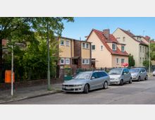 Wohnstraße mit einer Reihe von Einfamilienhäusern, teils aus gelbem Putz und rotem Ziegeldach sowie Klinkerfassaden. Im Vordergrund sind parkende Autos, ein orangefarbener Briefkasten und Straßenschilder sichtbar, während dichte Bäume die Szenerie ergänzen.