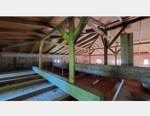 Dachboden mit sichtbarer Holzbalkenkonstruktion, Holzboden und freiliegenden Ziegelwänden. Kleine Fenster im Mauerwerk lassen gedämpftes Tageslicht herein.