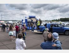 Eine Menschenmenge steht auf einem Parkplatz mit mehreren parkenden Autos im Hintergrund. Auf der rechten Seite ist ein blauer Pickup-Truck zu sehen, daneben ein blau-weißes aufblasbares Zelt mit der Aufschrift "In Märkisch [Land]". Der Himmel ist bewölkt, und die Umgebung wirkt wie ein offener Platz.