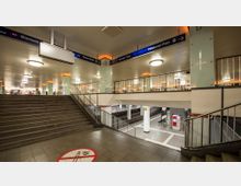 U-Bahn-Station mit Treppenaufgang, Geländern und einem Blick auf die Gleisebene. Wegweisschilder zeigen Richtungen zu Zielen wie "Potsdamer Platz" und "Leipziger Platz", und neonorange Beleuchtungen betonen die Decke und Wände.