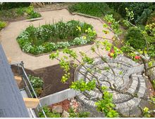 Blick auf einen Garten mit symmetrischen Wegen aus Kies, rechteckigen Beeten mit Blumen und Gräsern sowie einer runden Steinstruktur im Vordergrund. Ein blühender Baum mit roten Früchten ragt in das Bild hinein.