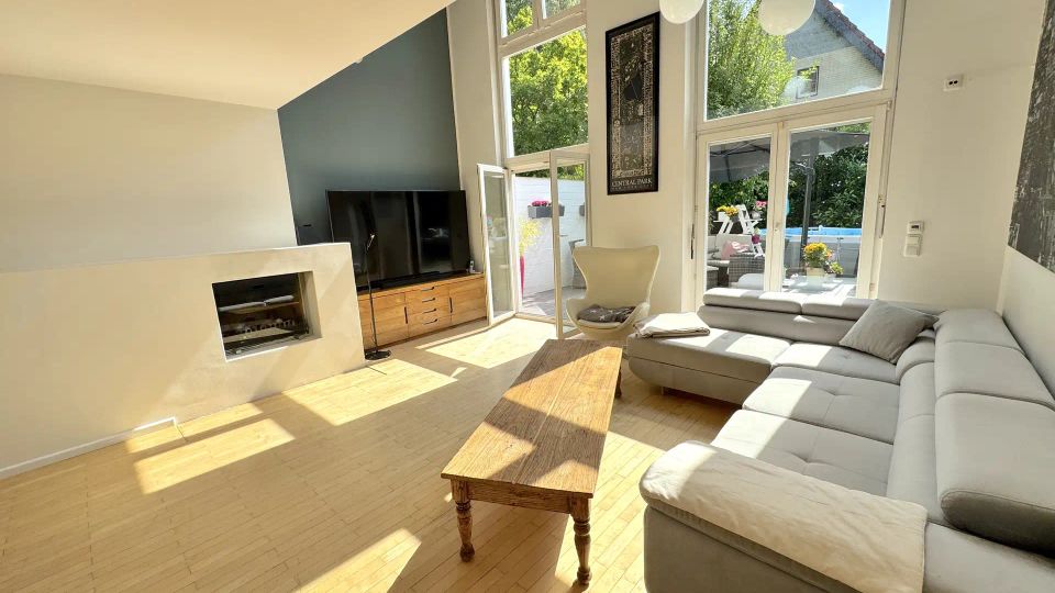Heller, moderner Wohnbereich mit hohen Fensterfronten und Terrassentür, die viel Tageslicht auf den hellen Holzboden lässt. Zu sehen sind ein L-förmiges Sofa, ein Holztisch, ein Kamin, ein Fernseher auf Sideboard sowie Blick in den begrünten Außenbereich.