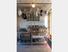 Industrieküche mit Edelstahlarbeitsfläche, einem großen Gasherd mit sechs Brennern und einem Ofen darunter. Oberhalb hängt an einem Metallgitter Kochgeschirr wie Töpfe, Pfannen und Kannen, während der Hintergrund aus Glasbausteinfeldern und einer Betonwand besteht.