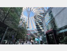 Moderne Architektur im Sony Center in Berlin, mit einem ikonischen Zeltdach, Glasfassaden und umliegenden Gebäuden. Im Vordergrund befinden sich Bäume, Geschäfte und ein Eingang zu einem Restaurant mit transparenten Terrassenüberdachungen.