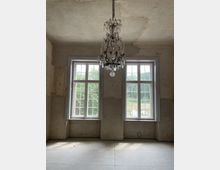 Ein leerer Raum mit zwei großen, weißen Sprossenfenstern, die Tageslicht hereinlassen. An der Decke hängt ein dekorativer Kronleuchter, und die Wände sind unverputzt mit sichtbaren Abnutzungsspuren.