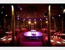 Innenansicht eines festlich dekorierten Zeltes mit runder Bühne in der Mitte, umgeben von runden Dinner-Tischen mit roten Tischdecken und eingedecktem Geschirr. Die Deckenstruktur ist kuppelförmig und mit gemustertem Stoff bespannt, während bunte Scheinwerfer den Raum beleuchten.