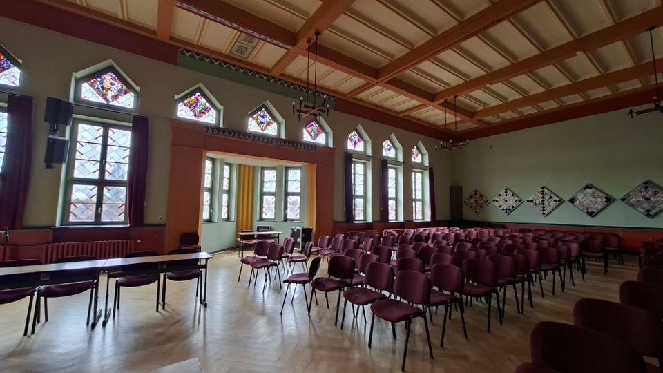 Innenraum eines Saals mit Parkettboden und vielen in Reihen aufgestellten bordeauxfarbenen Stühlen, seitlich ergänzt durch Tische. Hohe Fenster mit Buntglas-Oberlichtern und ein kassettierter Holzbalken‑Deckenaufbau prägen den Raum; an der rechten Wand hängen rautenförmige Wandtafeln.