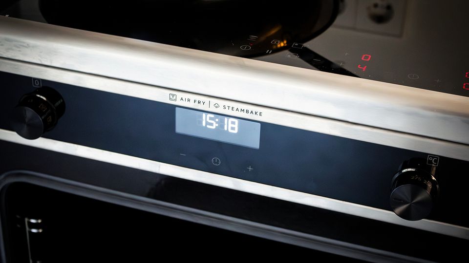 Nahaufnahme eines modernen Einbauherds mit schwarzer Glasfront und Edelstahlblende, mit zentralem Digitaldisplay und Drehknöpfen links und rechts. Oben ist ein Glaskeramik-Kochfeld mit leuchtenden Bedienanzeigen und einem Topf zu sehen.