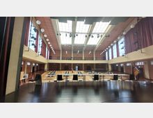 Großer Veranstaltungssaal mit hoher Decke und einer Glasdachkonstruktion, die viel Tageslicht hereinlässt. Der Raum ist mit Tischen und Stühlen ausgestattet, hat rote Vorhänge an den Seitenfenstern und ist mit Beleuchtungs- und Bühnentechnik versehen.