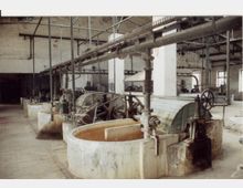 Industriehalle mit historischen Maschinen und großen runden Behältern aus Stein; darüber laufen Rohre und Metallstreben. Die Wände sind aus grobem Mauerwerk, und durch Fenster fällt Tageslicht in den Raum.