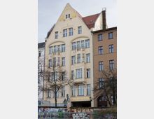 Mehrstöckiges helles Wohn- und Geschäftshaus mit geschwungenem Giebel und rotem Ziegeldach, vielen hohen Fenstern und kleinen Balkonen; im Erdgeschoss ein Durchgang mit der Aufschrift „Engelbecken-Hof“. Davor stehen kahle Bäume und eine niedrige Mauer mit Graffiti an einer Straßenkante.