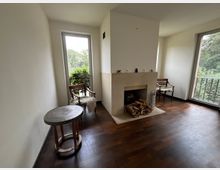 Ein helles Wohnzimmer mit einem Kamin in der Mitte, flankiert von zwei großen Fenstern, die den Blick ins Grüne ermöglichen. Der Raum ist mit dunklem Holzboden, zwei Stühlen und einem kleinen runden Tisch eingerichtet.