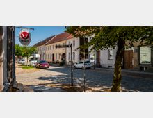 Blick auf eine gepflasterte Innenstadtstraße mit geparkten Autos, einem Straßenschild „Dammstraße“ und einer runden „Tierarzt“-Leuchtreklame am linken Gebäude. Im Hintergrund stehen zweigeschossige, helle Häuser mit roten Ziegeldächern und Café-Fassade, begleitet von Straßenbäumen am Rand.