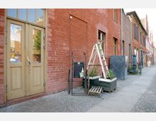 Straßenszene mit einer roten Backsteinfassade und großen Holztüren. Vor dem Gebäude stehen eine weiße Leiter, eine alte Sackkarre mit Werbung, Blumenkästen und eine Gehwegfläche aus Kopfsteinpflaster.