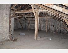 Ein leerer, verstaubter Dachboden mit sichtbaren Holzbalken, einem schrägen Dach und Wänden aus unverputztem Mauerwerk. Im Raum befinden sich vereinzelt Rohre, ein Plastikbehälter und verstreuter Unrat.