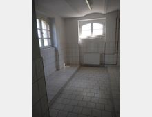 Ein gefliester Raum mit weißen Wand- und Bodenfliesen, einem erhöhten Podest an der linken Wand und zwei Fenstern mit Rundbögen. An der rechten Wand sind Heizungsrohre und ein Heizkörper sichtbar.