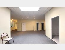 Ein einfacher Innenraum mit hellgelben Wänden, grauem Teppichboden und einer Rasterdecke mit eingebauten Leuchten. Im Hintergrund befinden sich drei Türen, eine mit einem Toilettenschild, und links ist ein großes Fenster mit Blick in einen angrenzenden Raum zu sehen.