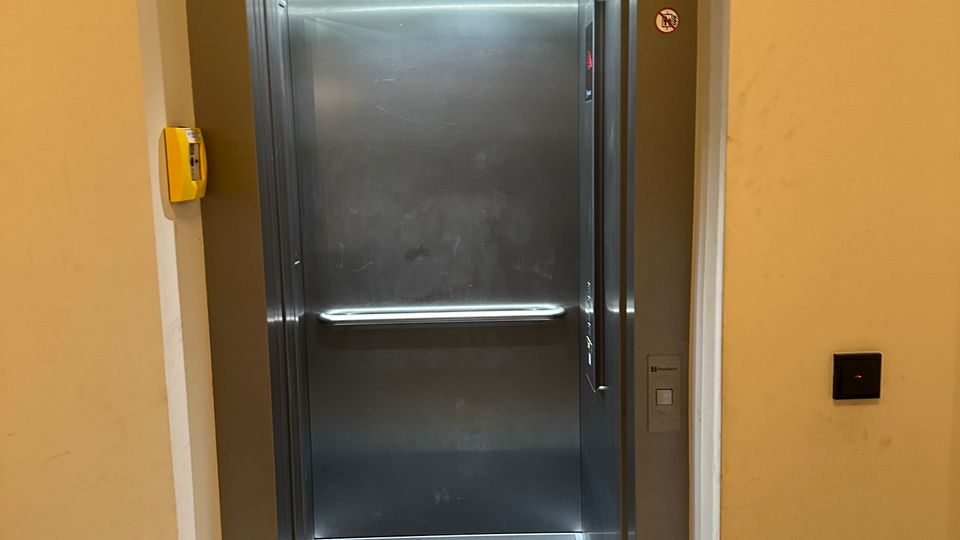 Offener Aufzug in einem Innenflur mit Edelstahlkabine, Deckenleuchten und seitlichem Bedienpanel. Der Aufzug ist in eine helle Wand eingelassen, mit Türrahmen aus Metall sowie Rufknopf und Hinweisschildern neben der Öffnung.
