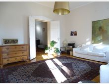 Wohnzimmer mit heller Einrichtung, darunter ein weißes Sofa, eine Holzkommode und ein großer orientalischer Teppich auf Parkettboden. Eine offene Doppeltür führt zu einem angrenzenden Raum; an den Wänden hängen Gemälde, und eine Zimmerpflanze steht im Sonnenlicht.