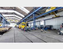 Innenansicht einer großen Industriehalle mit hohen Decken und Stahlkonstruktionen. Die Halle enthält Schienenfahrzeuge, Werkbänke, Maschinen und ein gelbes Wartungsfahrzeug, während Schienen den Boden durchziehen.